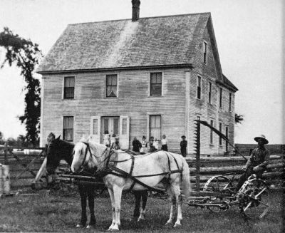 two horses pulling a mower in front of a 2 story clapboard house