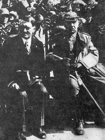 Hindenburg in full uniform sitting next to Hitler in front of a crowd