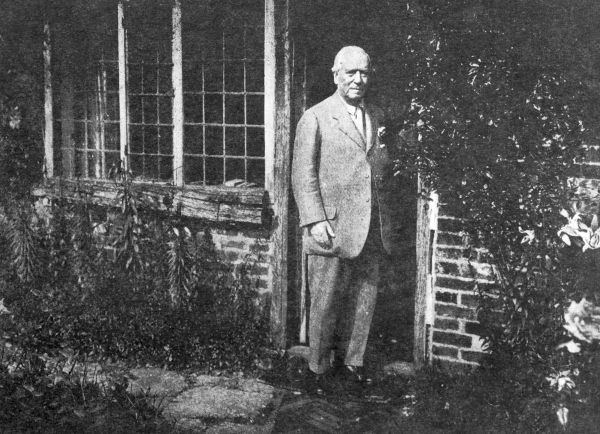 white-haired man in a suit standing in the doorway of a cottage