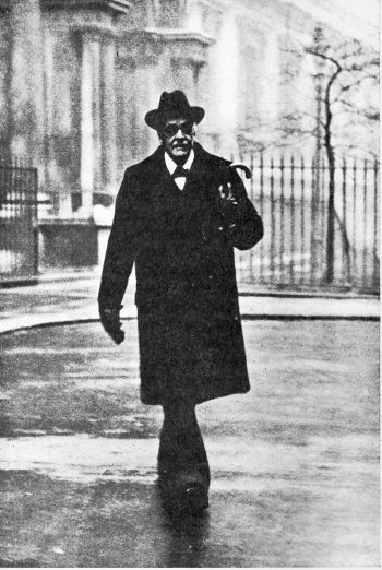 man in long black coat, hat and sunglasses walking in front of building