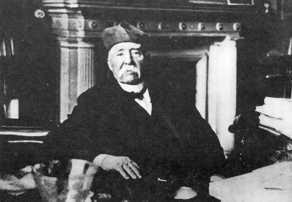 man with white droopy moustache sitting at a desk in front of fireplace