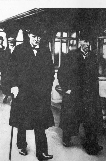 2 men walking on a street in long coats, bowler hats, young Churchill with a cane