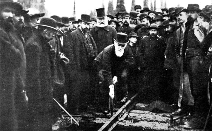 crowd watching white-bearded man in top hat drive a railway spike