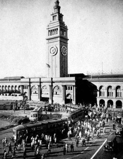 Two story building with clock tower and trams in front unloading many people