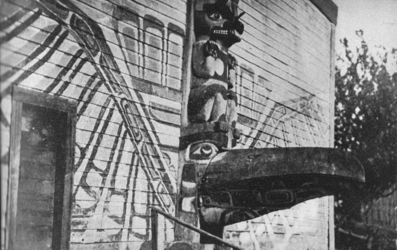 Photo of the outside of a indigenous community house with a totem pole in front of it.