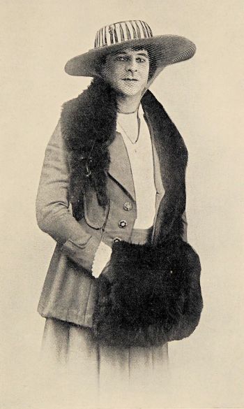 author wearing a woman’s suit with large hat with a mink around neck and a mink hand muffler