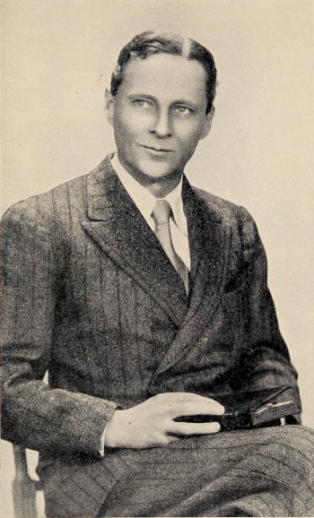 author seated with book wearing a striped suit and tie