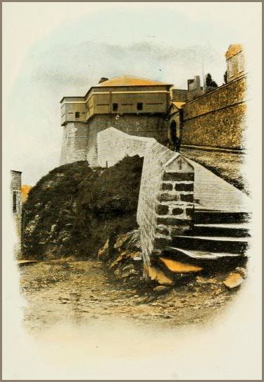 Image shows a stone staircase climbing toward a squat, thick-walled hilltop fort, its small windows and heavy masonry rising from bare rock and earth.