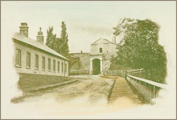 Image shows a dirt road leading to a massive stone gate, flanked by a long row of low buildings on the left and a raised wooden walkway on the right. In the background can be seen trees and other buildings.