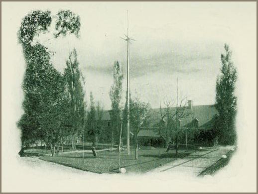 Image of a courtyard with a central flagpole, tree-lined paths, and a large building in the background.