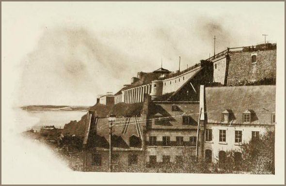 Image of a fortified hilltop structure with stone walls and sloped roofs overlooking a body of water.